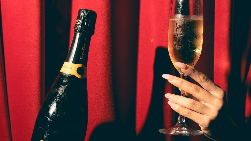 A hand with manicured nails holds a glass of champagne next to an open bottle, set against a bright red curtain—capturing the glamour and intrigue found Behind the Red Door. Lifestyle