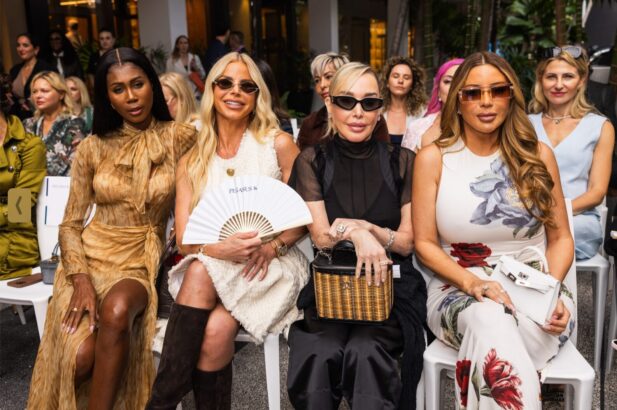 Four stylish women sit in the front row at a fashion show, dressed in elegant outfits and sunglasses, holding programs and accessories, embodying the latest fashion trends as an outdoor audience looks on. Lifestyle