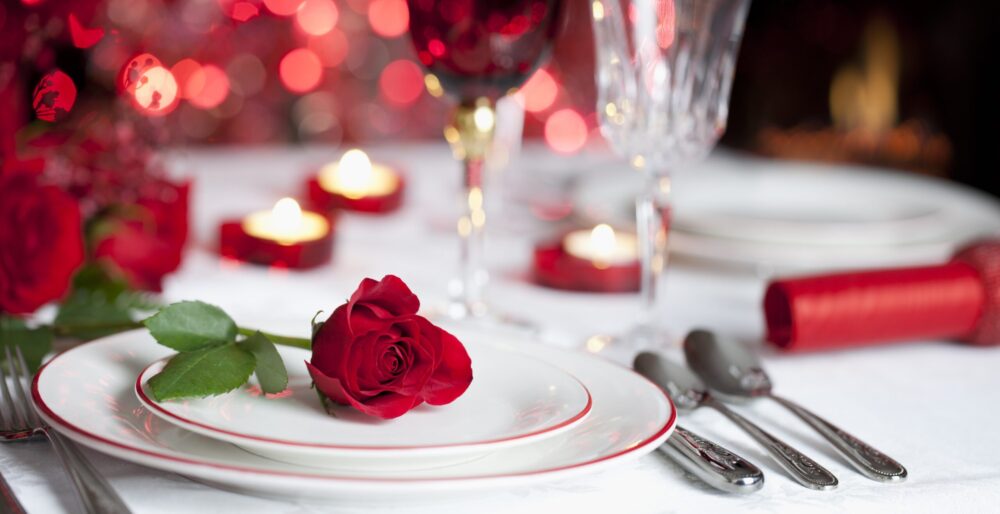 A romantic table setting with a single red rose on a white plate, surrounded by cutlery, wine glasses, and lit candles, creates a warm atmosphere filled with hearts and red bokeh lights in the background. Lifestyle