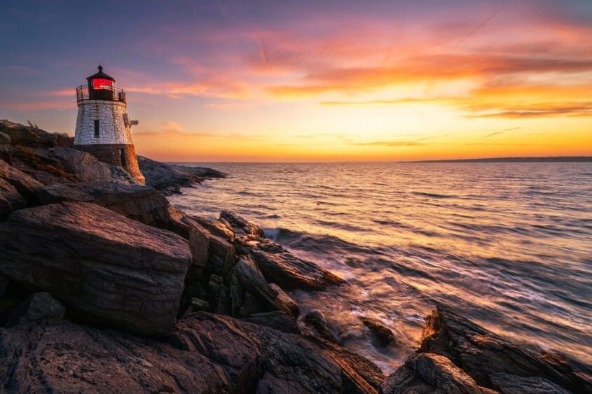 A lighthouse with a glowing red light stands on rocky cliffs overlooking a calm ocean at sunset—an idyllic scene for Romantic Escapes, with vibrant orange, yellow, and purple hues filling the sky and reflecting on the water. Lifestyle