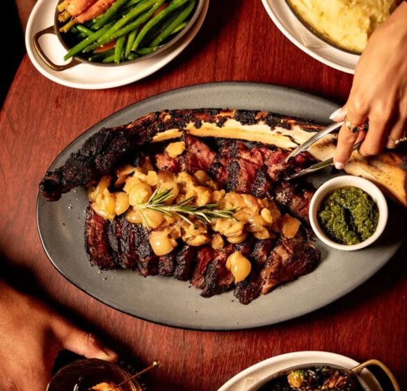 A sliced, bone-in steak topped with garlic and rosemary sits on a platter with a side of green sauce. Plates of green beans, mashed potatoes, and hands reaching for food radiate the heat and hedonism of a lively meal around a wooden table. Lifestyle