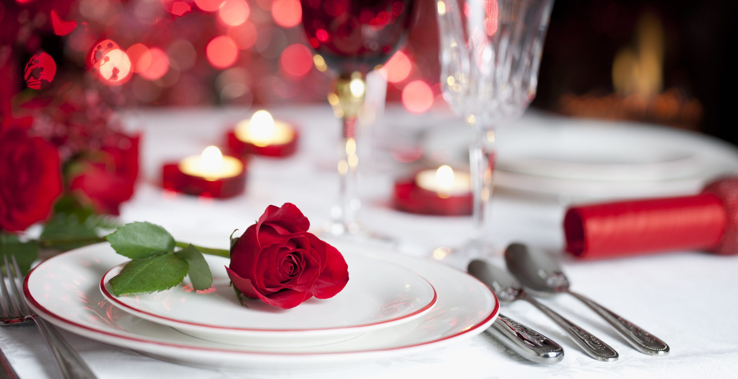A romantic table setting with a single red rose on a white plate, surrounded by cutlery, wine glasses, and lit candles, creates a warm atmosphere filled with hearts and red bokeh lights in the background. Lifestyle