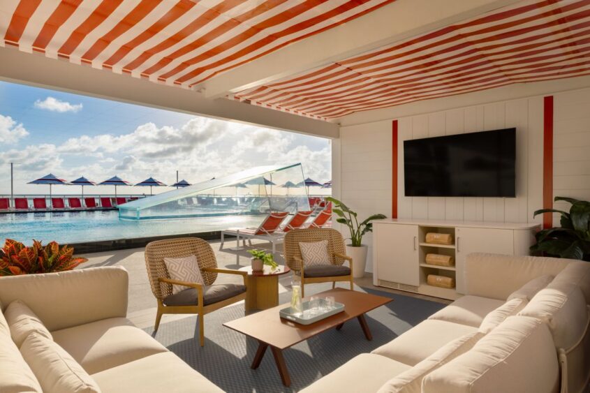 A stylish poolside cabana in Fort Lauderdale with modern beige sofas, a coffee table, and a TV. Outside, a pool with a waterslide and orange lounge chairs is visible under blue sky and striped umbrellas—perfect for a Valentine’s getaway. Lifestyle