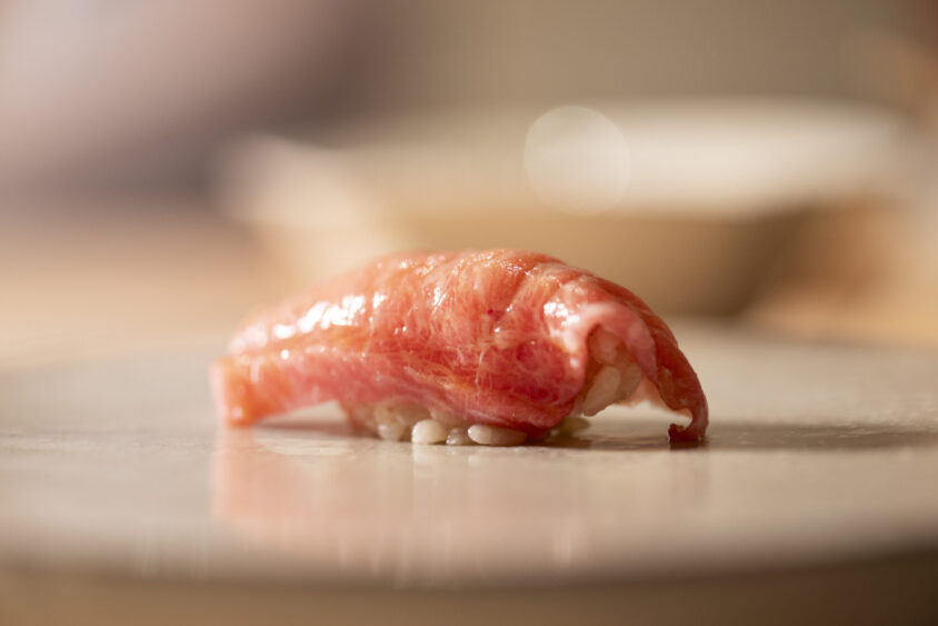 A piece of nigiri sushi with a slice of marbled raw fish placed over a small mound of white rice, served on a light-colored plate—reflecting centuries of craft in every detail, against a softly blurred background. Lifestyle