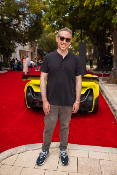 A man wearing sunglasses, a black shirt, and gray pants stands smiling on a red carpet outdoors, embodying the spirit of Driven By Design, with a yellow sports car and trees in the background. Lifestyle