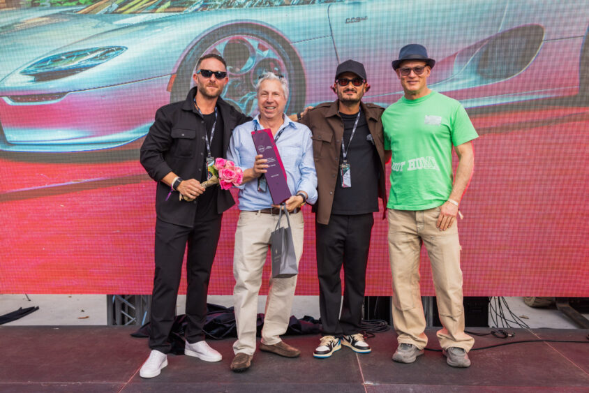 Four men stand side by side on a stage, posing for a photo. One holds a bouquet, another holds a red box and bag. A "Driven By Design" sports car image is displayed on the large screen behind them. Lifestyle