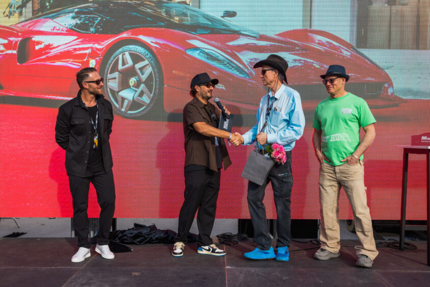 Four men stand onstage in front of a large screen displaying a red sports car; two men in the center shake hands and smile while the others look on, celebrating innovation that's truly Driven By Design. Lifestyle