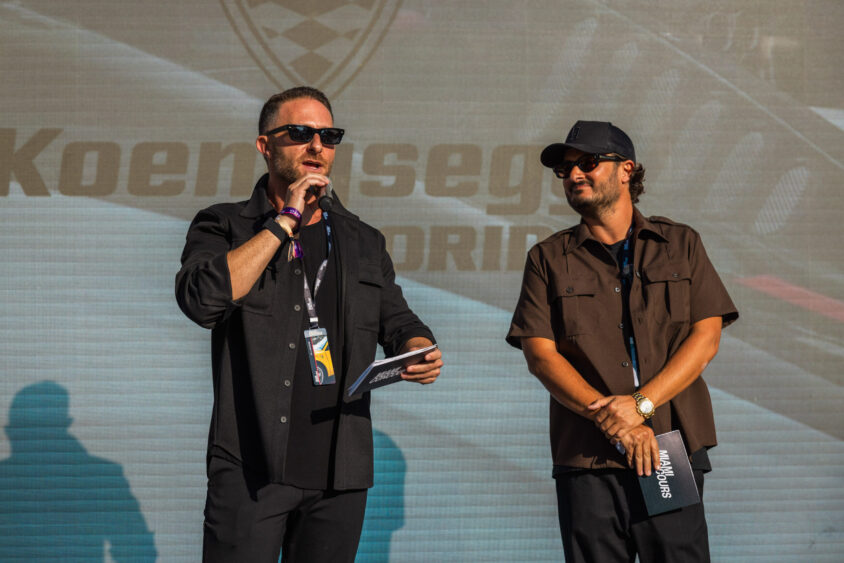 Two men stand on stage in front of a Koenigsegg backdrop, holding microphones and cards. Both are dressed in dark casual clothing and sunglasses, presenting at an event that's clearly Driven By Design. Lifestyle