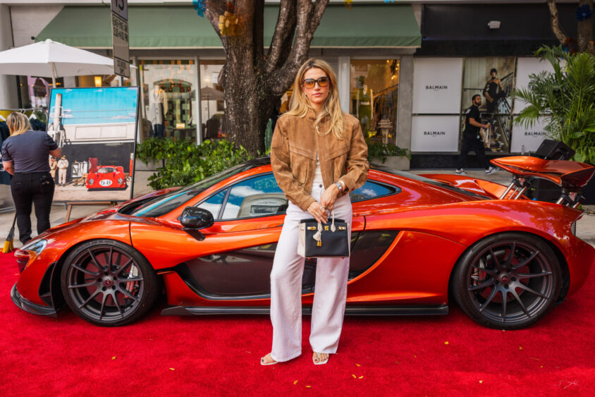 A stylish woman in sunglasses, a brown jacket, and white pants stands on a red carpet before an orange sports car by upscale shops. Driven By Design, she holds a black handbag and looks confidently at the camera. Lifestyle