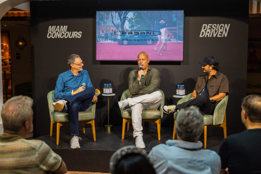 Three men sit on stage in a panel discussion at the Miami Concours event. They hold microphones as the audience watches. Behind them, a screen displays "Pagani Automobili," “Miami Concours,” and the phrase "Driven By Design. Lifestyle