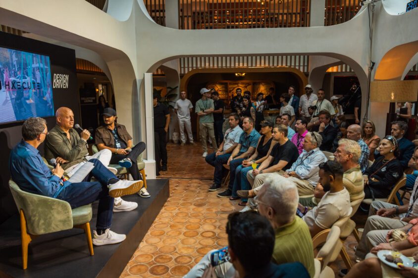 A panel of three people speaks to a seated audience in a modern, stylish room, with a large screen displaying "DRIVEN BY EXECUTION" and "Driven By Design" in the background. The audience listens attentively. Lifestyle
