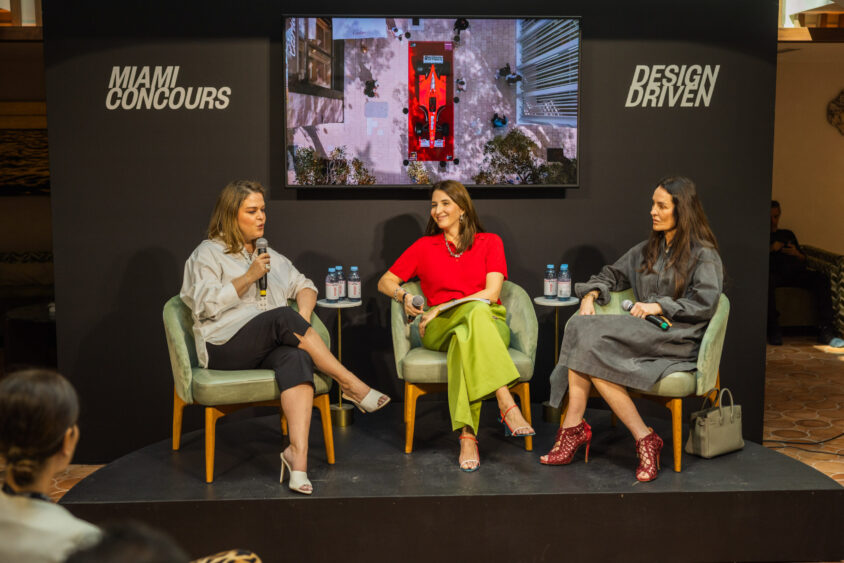 Three women sit on stage in armchairs, engaged in a Driven By Design panel discussion at the Miami Concours event. Behind them, a screen displays an overhead car image and event branding. Bottled water rests on a small table beside them. Lifestyle