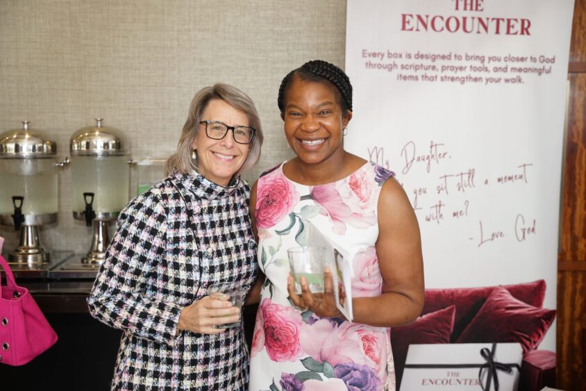 Two women smiling and standing together at a 4KIDS Broward Galentine’s fundraising event. One wears glasses and a checkered dress; the other wears a white dress with pink flowers and holds a drink. A banner and beverage dispensers are visible behind them. Lifestyle