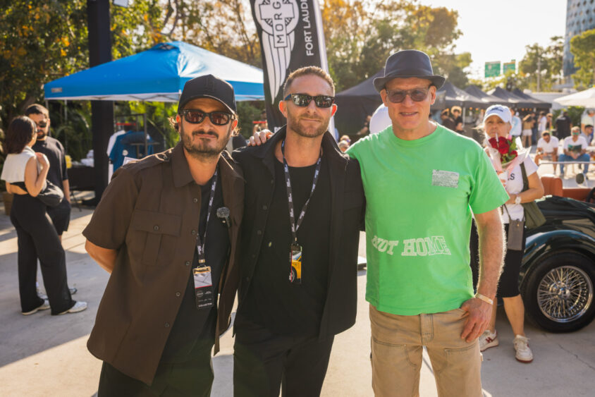 Three men pose together outdoors at a Driven By Design event, with two in dark shirts and sunglasses, and one in a green t-shirt and hat. In the sunny background, tents, people, and a classic car add to the lively scene. Lifestyle
