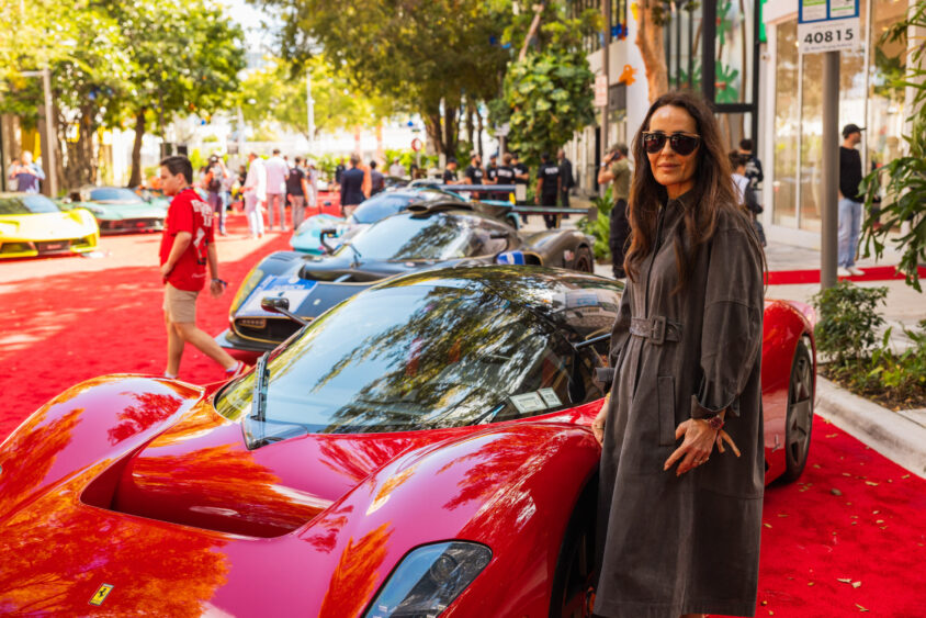 A woman in a long coat and sunglasses stands next to a shiny red sports car at an outdoor car show, surrounded by other luxury cars and people—an event truly Driven By Design. Lifestyle