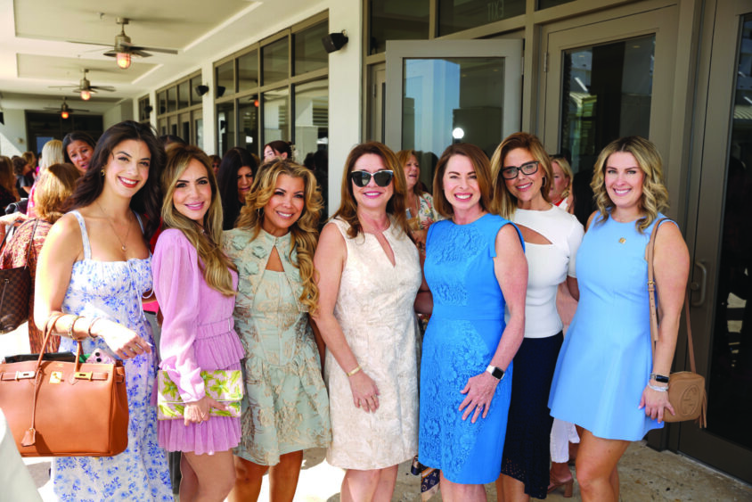 A group of seven women dressed in colorful spring and summer outfits smile and pose together outside a modern building with glass doors at a Broward Galentine’s fundraising social gathering. Lifestyle
