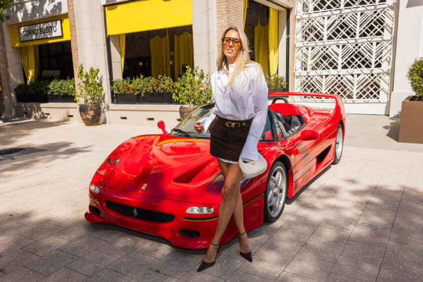 A woman in sunglasses, a white blouse, and a brown skirt poses smiling in front of a red Ferrari sports car—Driven By Design—parked on a sunny sidewalk outside a building with yellow awnings. Lifestyle