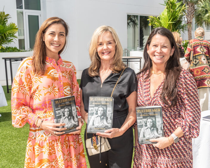 Three women stand outdoors, smiling and holding copies of the book "Rulebreaker" at a Big Ideas event. They are dressed in colorful, patterned dresses, with greenery and a modern building in the background. Lifestyle