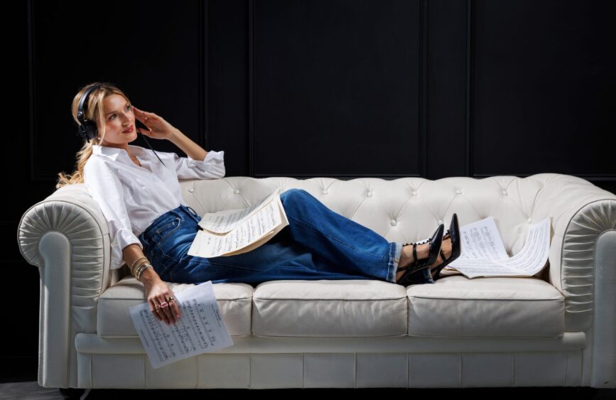 A woman wearing headphones and casual clothes lounges on a white sofa, studying sheet music with bass lines highlighted, more sheets on her lap and scattered around her, against a dark background. Lifestyle