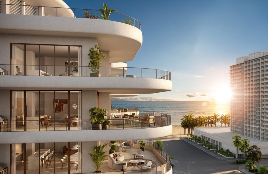 Modern beachfront apartment building with large balconies and glass railings, overlooking the ocean and palm trees at sunset. The sky is clear, and sunlight reflects off the water and nearby boutique hotel buildings. Lifestyle