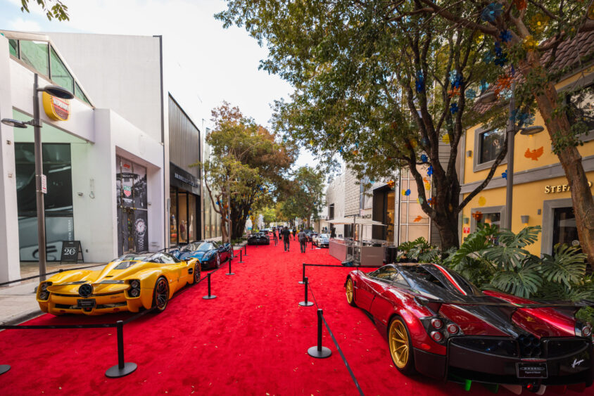 A row of luxury sports cars, driven by design, is displayed on a bright red carpet along a street lined with shops and trees, as people walk by and admire the stunning vehicles in the background. Lifestyle