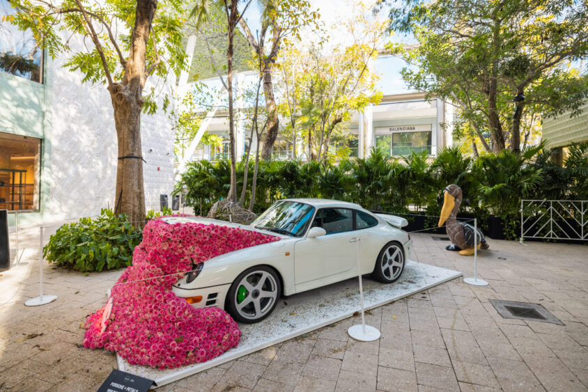 A white sports car, Driven By Design, is displayed outdoors, half-covered in a dense cascade of bright pink flowers. Surrounded by greenery and trees, the scene includes upscale shop buildings and a Balenciaga storefront in the background. Lifestyle