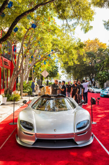 A sleek silver sports car, driven by design, is displayed on a red carpet outdoors, surrounded by trees and people admiring and photographing it on a sunny day. Lifestyle