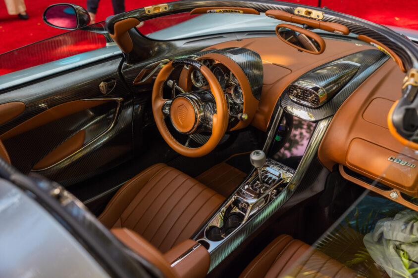 Close-up view of a luxury sports car interior, Driven By Design, featuring brown leather seats, carbon fiber details, a round steering wheel, manual gear shifter, and modern dashboard, all visible with the convertible top down. Lifestyle