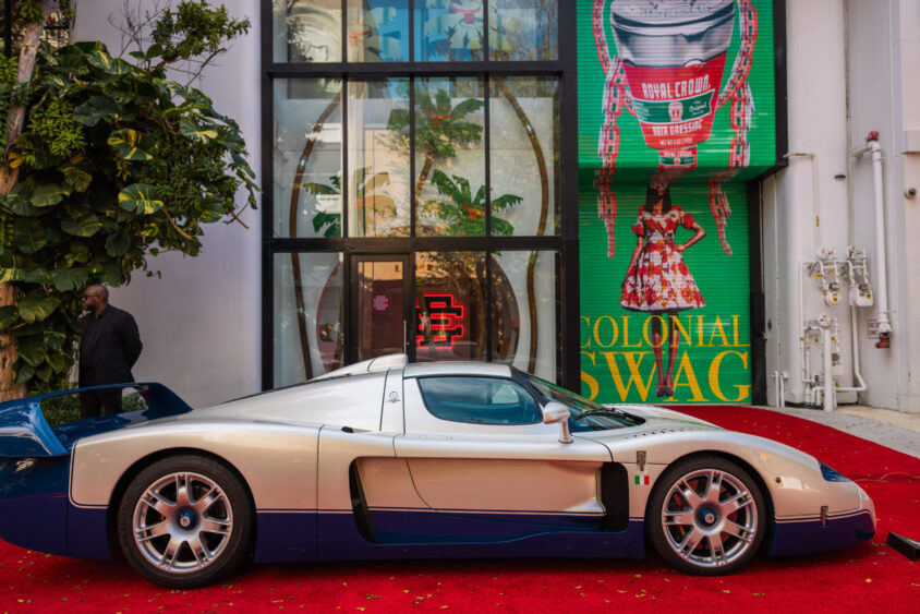 A silver and blue sports car, Driven By Design, is parked on a red carpet in front of a building with glass doors and a colorful mural featuring a person in plaid attire, a large can, and the words "Colonial Swag. Lifestyle