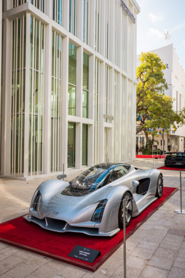 A sleek silver hypercar, driven by design, is displayed on a red carpet outside a modern glass building, surrounded by ropes. Sunlight highlights the car's aerodynamic form and sharp edges. Lifestyle