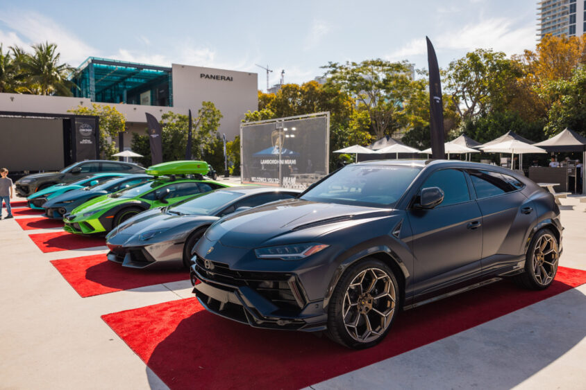 A row of luxury Lamborghini cars, driven by design, including SUVs and sports cars, are displayed on red carpets outdoors at an auto event, with trees, tents, and modern buildings in the background under a sunny sky. Lifestyle