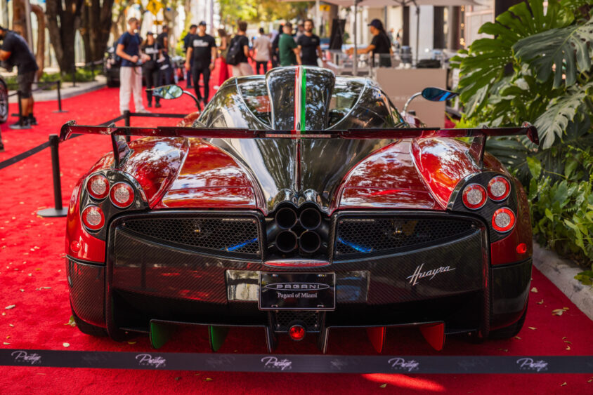 A red and black Pagani Huayra supercar, Driven By Design, is displayed on a red carpet outdoors, surrounded by black velvet ropes, with people and greenery in the background. Lifestyle