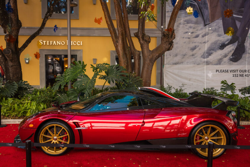 A shiny red sports car with gold wheels is displayed on a red carpet outdoors, surrounded by plants and trees, in front of a Stefano Ricci store—an impressive scene truly Driven By Design. Lifestyle