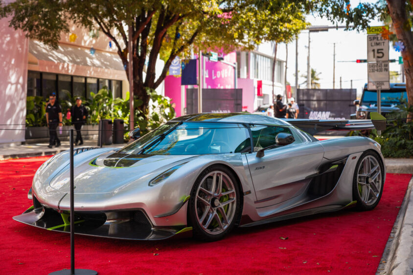 A sleek silver sports car with green accents, driven by design, is displayed on a red carpet outdoors, surrounded by trees, city buildings, and street signs. The car features a low, aerodynamic profile and large wheels. Lifestyle