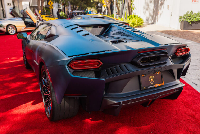 A matte blue Lamborghini sports car, driven by design, is parked on a red carpet outdoors with its rear facing the camera. Other luxury cars and palm trees can be seen in the background on a sunny day. Lifestyle