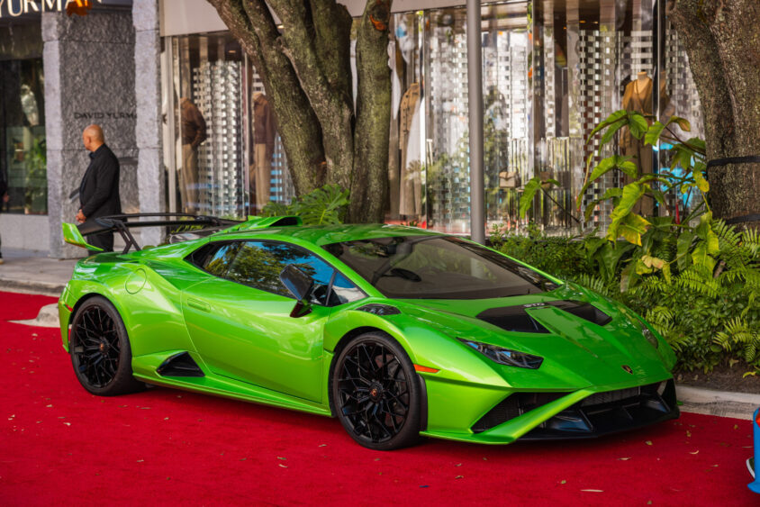 A bright green Lamborghini sports car, driven by design, is parked on a red carpet near a luxury storefront, with greenery and trees nearby and a person standing in the background. Lifestyle
