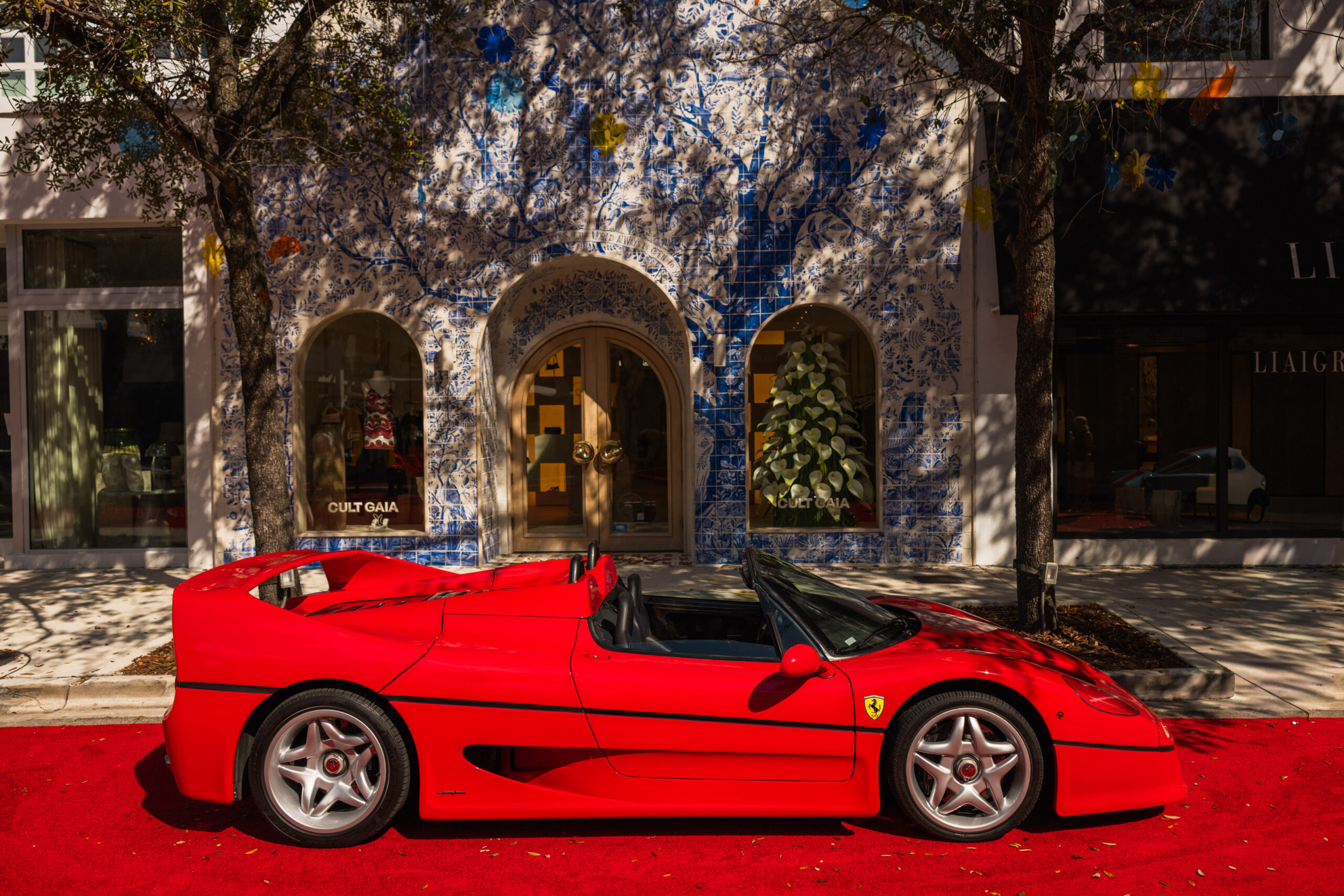 A red Ferrari convertible sports car, driven by design, is parked on a red carpet in front of a building adorned with blue and white mosaic wall art and framed by large trees casting shadows. Lifestyle