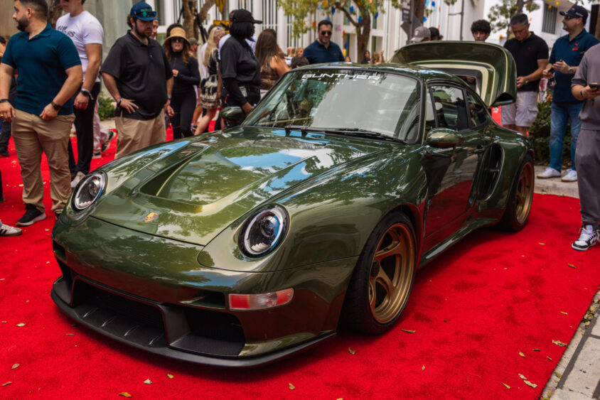 A dark green custom Porsche sports car with gold rims, driven by design, is displayed on a red carpet at an outdoor car show, surrounded by people taking photos and admiring the vehicle. Lifestyle