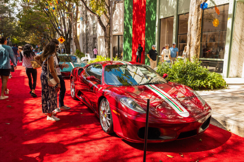 A shiny red Ferrari sports car with green and white racing stripes, driven by design, is parked on a red carpet outdoors, surrounded by people admiring it. Trees and shops line the street under sunny weather. Lifestyle
