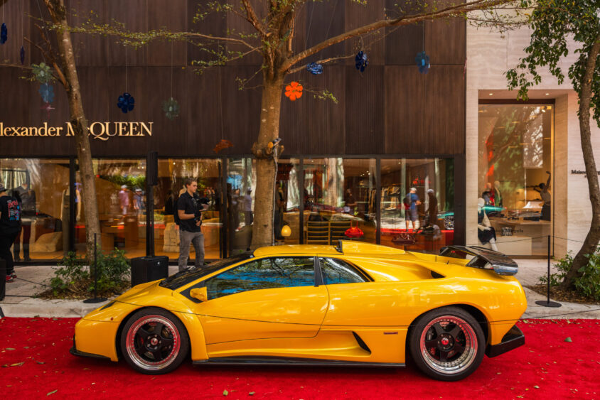 A yellow Lamborghini sports car, Driven By Design, is parked on a red carpet in front of an Alexander McQueen store, with people walking by modern glass storefronts and trees overhead. Lifestyle
