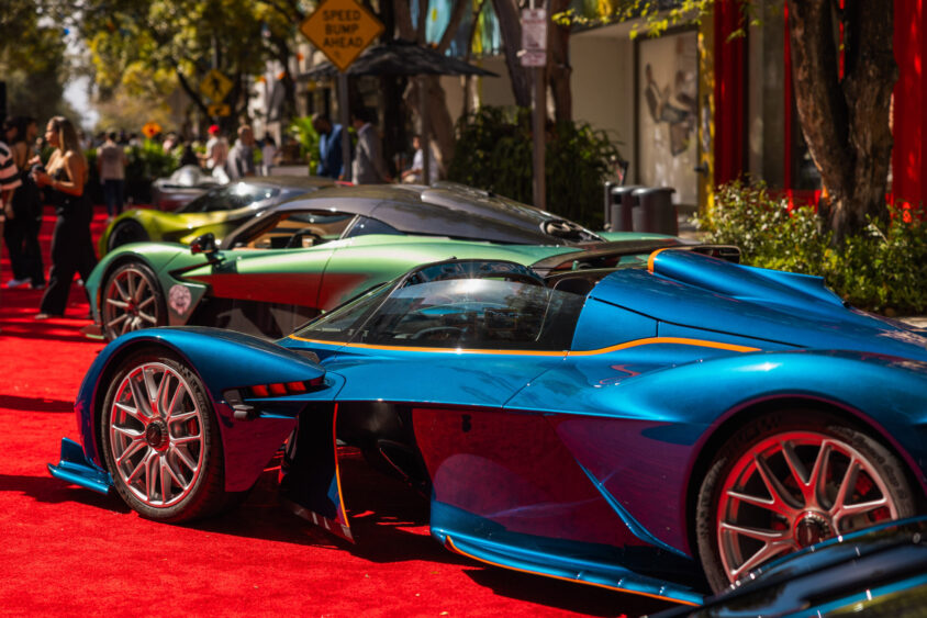 Three colorful luxury sports cars, including a blue one in the foreground, are parked on a red carpet outdoors, showcasing a scene truly driven by design, with trees and people in the background under sunny weather. Lifestyle