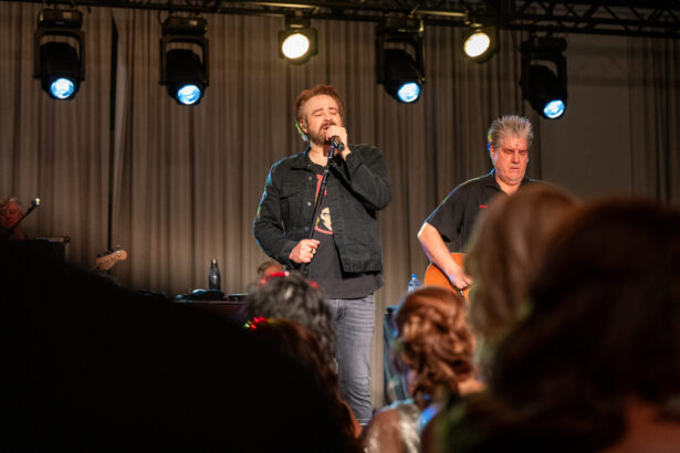 A male singer performs on stage holding a microphone, with a guitarist playing beside him. Stage lights shine overhead as an engaged community audience watches from the foreground. Lifestyle