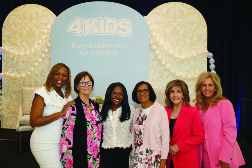 Six women in professional attire smile in front of an ornate “4KIDS. Every child deserves a seat at the table” backdrop, celebrating Broward Galentine’s and supporting 4KIDS fundraising efforts. Lifestyle