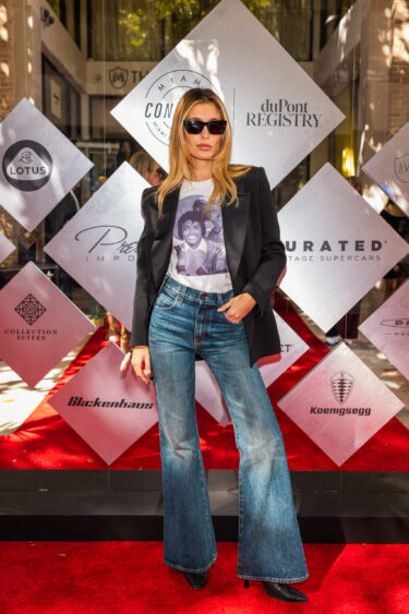A woman wearing sunglasses, a black blazer, a graphic T-shirt, and flared blue jeans poses on the red carpet in front of a backdrop featuring various logos and diamond-shaped panels, embodying a style that's truly Driven By Design. Lifestyle