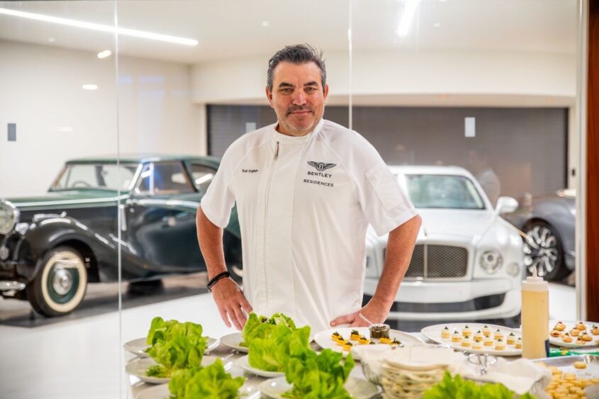 A chef in a white Bentley-branded uniform stands behind a table with plated appetizers and lettuce, with two luxury cars visible in the background inside The House That Bentley Built, a bright, modern garage. Lifestyle