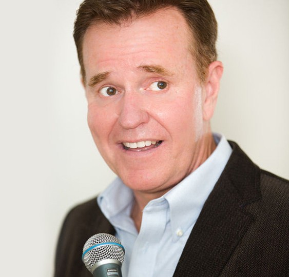 A man in a suit jacket and light blue shirt holds a microphone and smiles while looking slightly to the side, as if ready to host Two Nights, Three Shows, standing against a plain, light background. Lifestyle