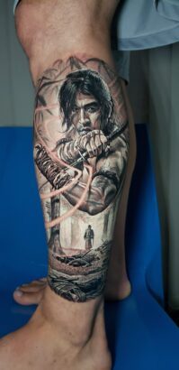 A Flesh and Canvas masterpiece, this realistic calf tattoo depicts a long-haired warrior drawing a bow, another figure in the background, and a rocky landscape—all brought to life with intricate shading and detailed artwork. Lifestyle