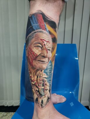 A detailed tattoo from Flesh and Canvas shows an elderly Indigenous person wearing traditional jewelry and a feathered headdress on a lower leg, displayed while seated on a blue chair. Lifestyle