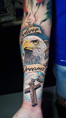 A detailed forearm tattoo on the canvas of flesh features a bald eagle, rosary with crucifix, and banners reading "Believe your Dreams," all set against a background of soft colors. Lifestyle