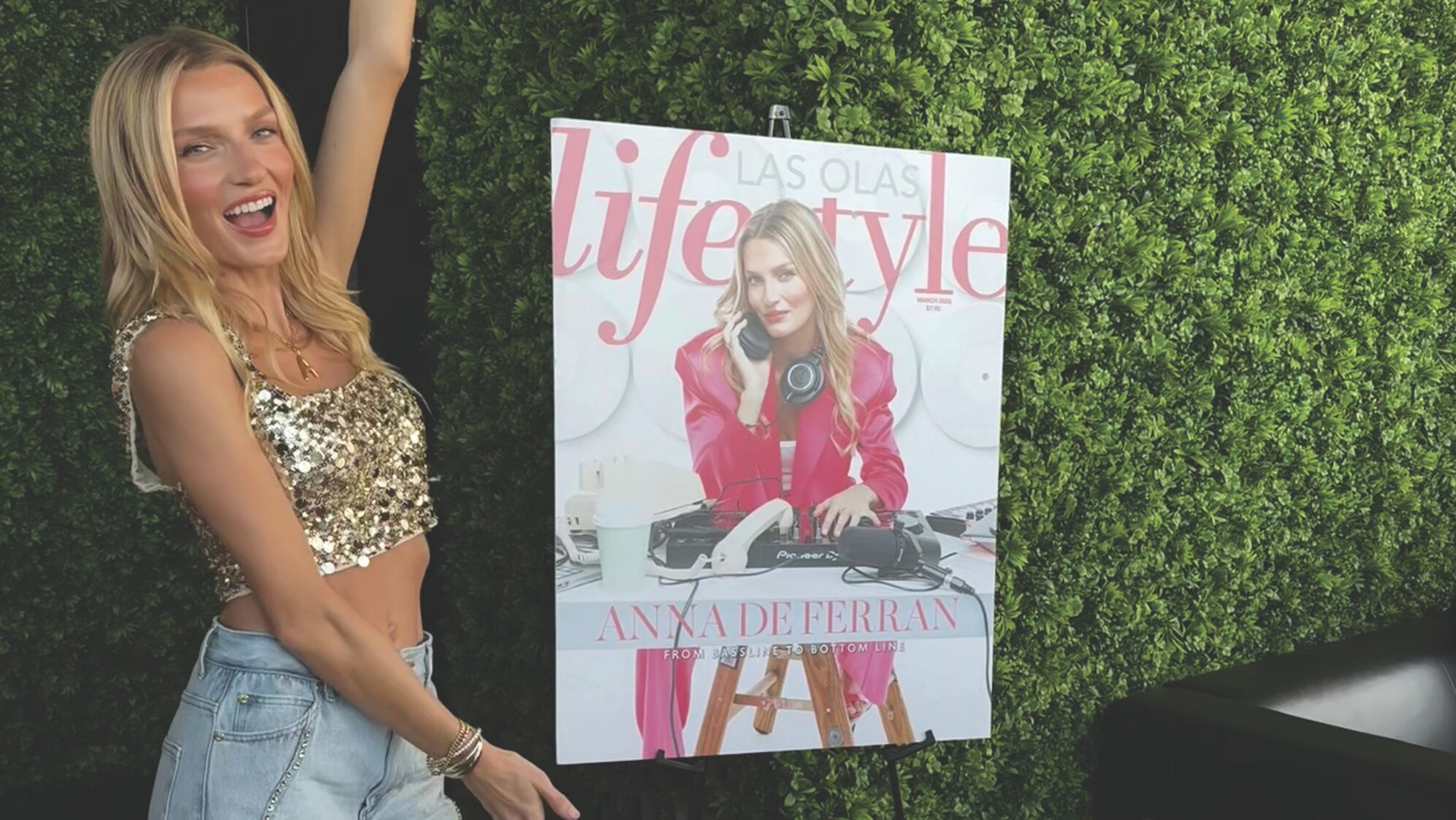 A smiling woman in a gold sequin crop top and denim shorts poses with one arm raised next to a large Las Olas Lifestyle March Cover Party magazine cover. The scene is set against a green leafy wall at Palm Garden Event House. Lifestyle